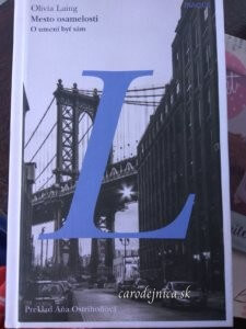Titulka knihy Mesto osamelosti s veľkým modrým písmenom L a na pozadí most nad ulicou a budovou